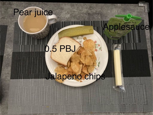 A cup of pear juice sits just above and to the left of a small plate of food consisting of half of a peanut butter and jelly, a handful of jalapeno chips, and a pickle. To the right of the plate are a stick of string cheese and a squeeze pouch of applesauce.