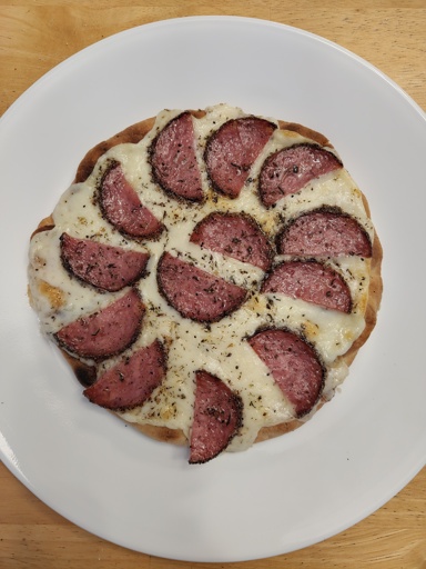Top - down view of a small round pizza with a thin golden - brown crust, topped with melted white cheese, evenly spaced slices of reddish - brown cured meat (likely salami), and sprinkled herbs. The pizza rests on a white plate placed on a light wooden surface.